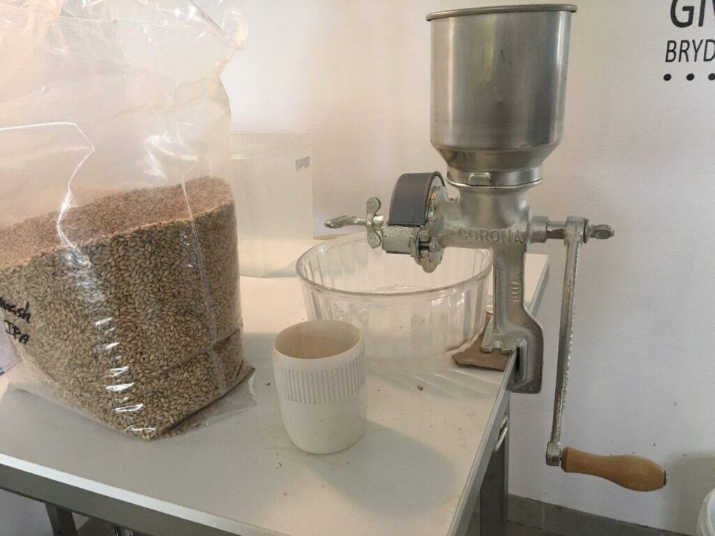 Pose med malt ved siden af en håndtrukket maltmølle og en skål på et bord